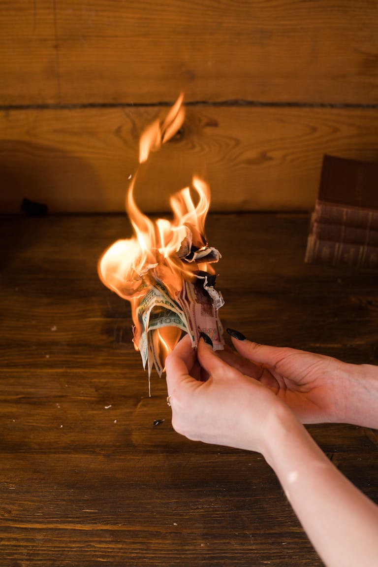 Hands holding burning banknotes against a wooden background, symbolizing financial waste.
